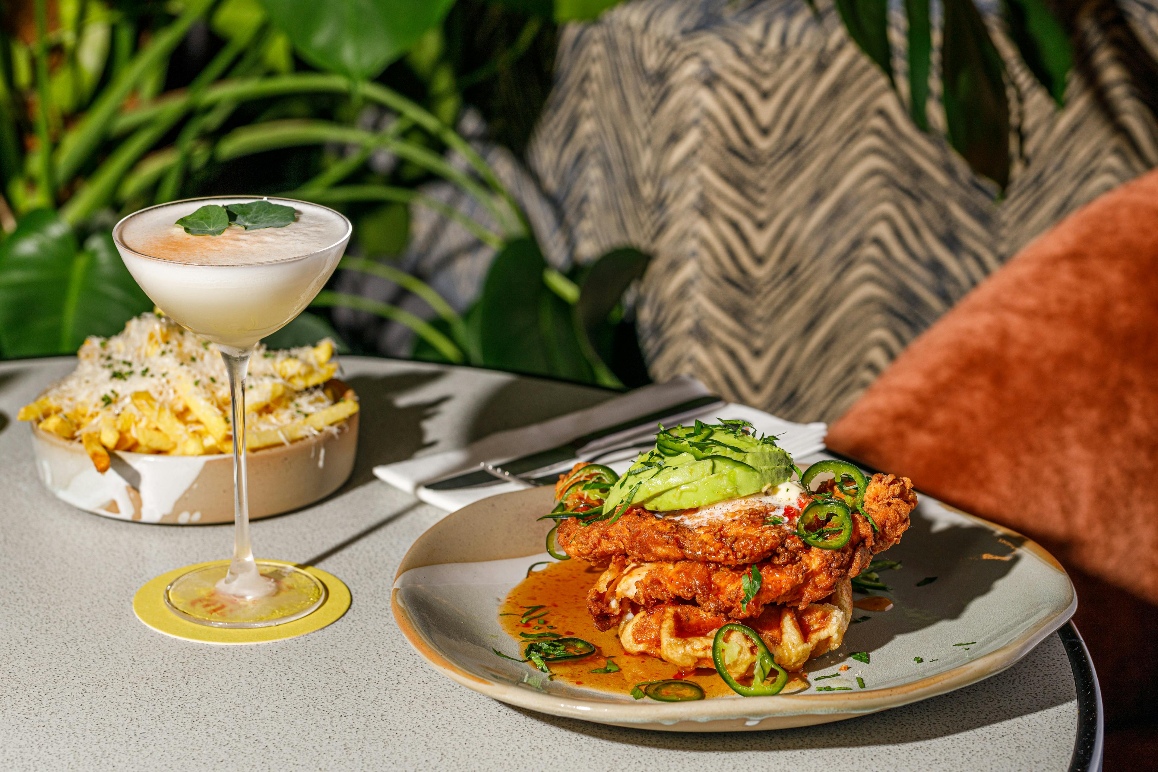 Cocktail beside fried chicken on waffles with cheesy chips behind on an outdoor table with foliage in the background.